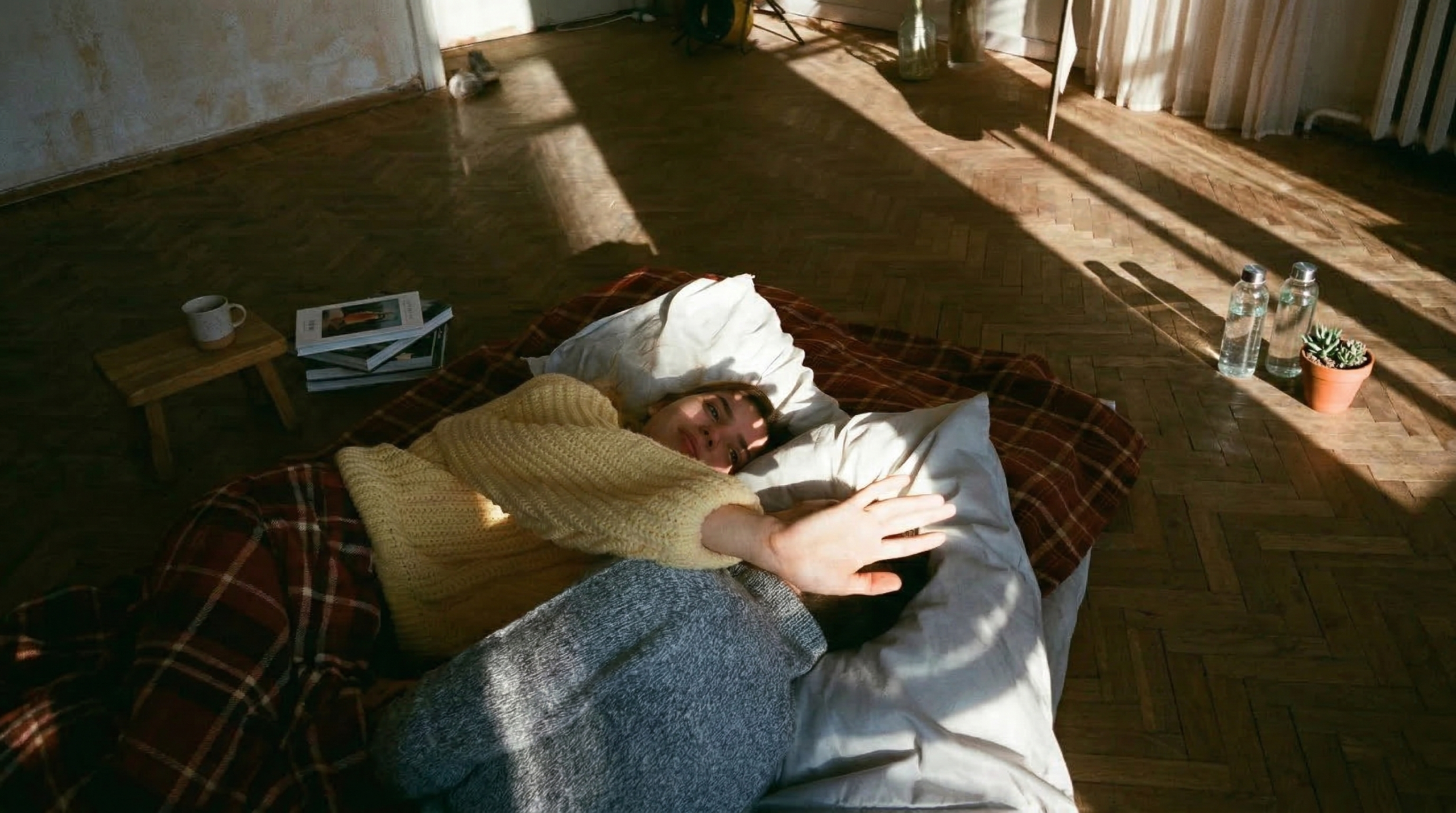 Une vue en plongée de deux personnes allongées confortablement sur un matelas posé à même un parquet ancien en chevrons. La pièce est baignée d'une lumière matinale chaleureuse qui crée des jeux d'ombres allongées. Une personne porte un pull en maille jaune et lève la main pour parer les rayons du soleil, tandis que l'autre, en pull gris, est vue de dos. Autour d'eux, une ambiance bohème avec quelques livres empilés, une tasse de café sur un petit tabouret en bois, des bouteilles d'eau et une petite plante en pot.