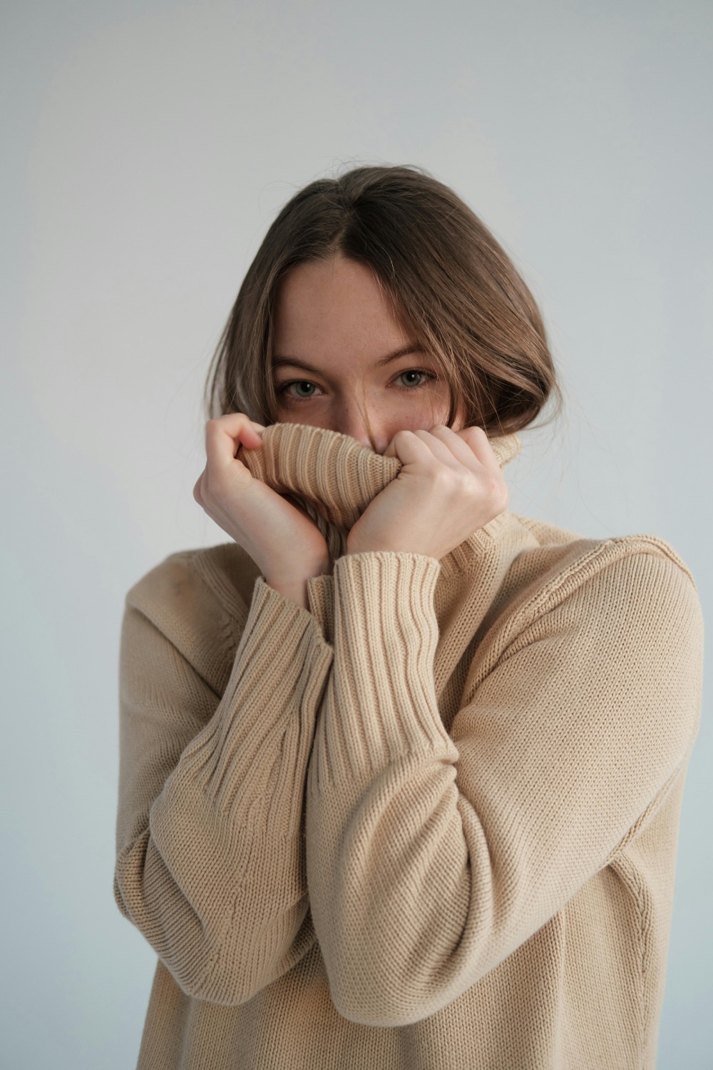 Un portrait rapproché en studio d'une femme aux cheveux longs et clairs, qui cache la moitié de son visage en relevant le col montant nervuré de son pull en tricot côtelé de couleur beige sable. Ses yeux clairs sont les seuls éléments du visage visibles, regardant directement l'objectif avec intensité. Ses mains, avec des ongles naturels, maintiennent fermement le tissu du col. L'éclairage est doux et direct sur un fond blanc propre.