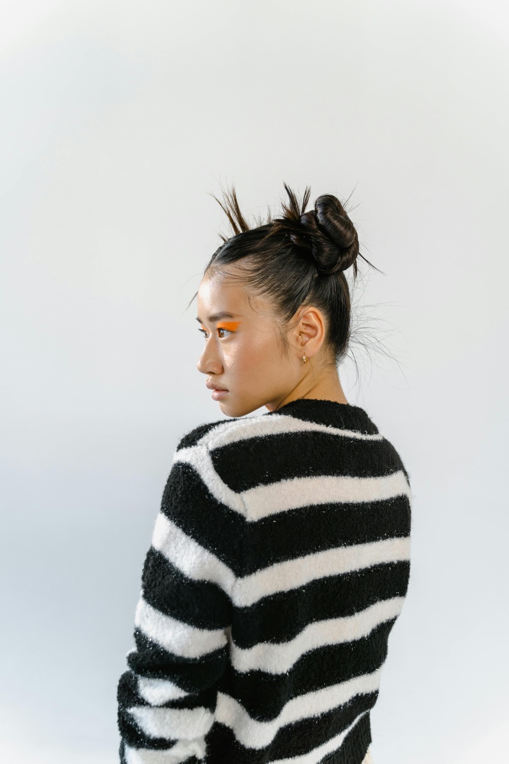 Un portrait photographique stylisé de profil arrière et latéral d'une femme asiatique avec une coiffure élaborée : ses cheveux noirs sont tirés vers le haut en une série de petits chignons serrés et de pointes hérissées. Elle regarde vers la gauche, montrant son profil. Son maquillage est dramatique, avec une large bande d'ombre à paupières orange vif qui s'étend sur le côté de son visage. Elle porte un pull en tricot flou à rayures horizontales noires et blanches audacieuses. Sa boucle d'oreille en or est visible. L'arrière-plan est un studio blanc minimaliste et uni. L'éclairage est vif et propre.